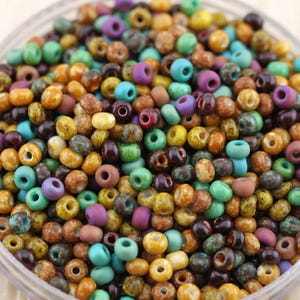 May include: A close-up of a bowl filled with small, round beads in various colours. The beads are teal, purple, brown, yellow, and orange. These beads are likely used for jewellery making or other craft projects.