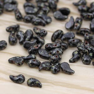 May include: A collection of black and green paisley-shaped glass beads with a speckled pattern. The beads are scattered on a white fabric surface.