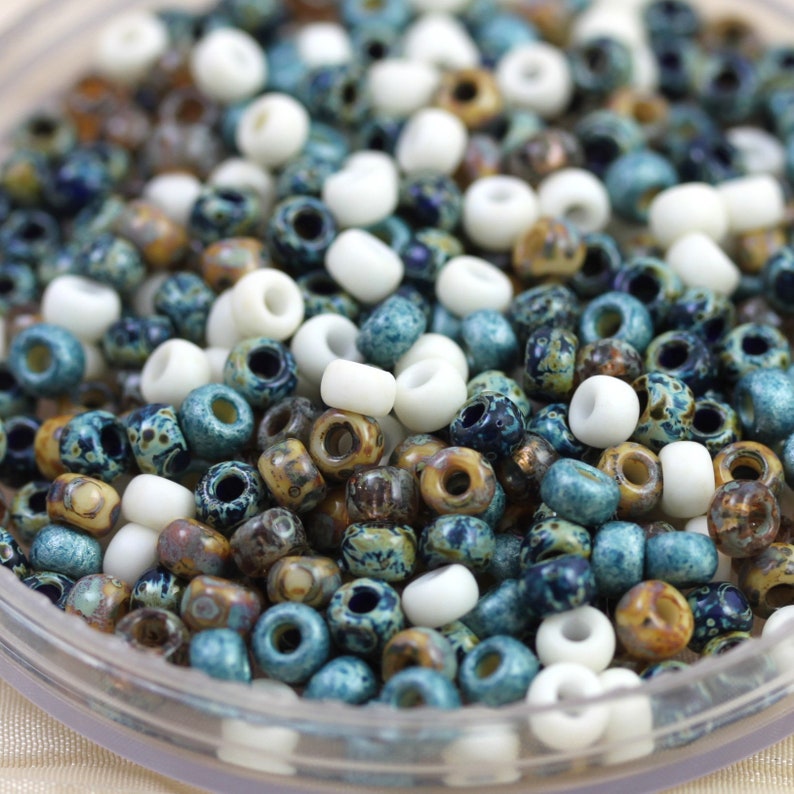 May include: A close-up of a variety of small, round beads in white, brown, and blue colors. The beads are in a clear container and appear to be made of glass or ceramic.