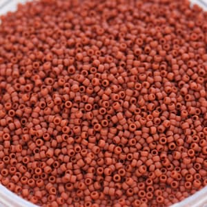Pode incluir: Uma visão de perto de uma grande quantidade de pequenas contas redondas de cor terracota. As contas são uniformes em tamanho e forma, e estão dispostas em um padrão aleatório.