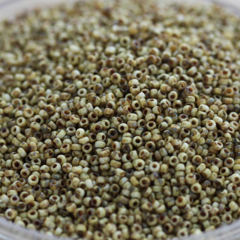 May include: A close-up of a large quantity of small, round, white beads with brown speckles. The beads are arranged in a random pattern and appear to be made of glass or ceramic.