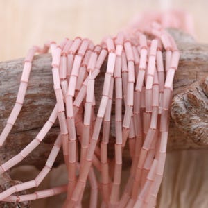 May include: Close-up of strands of light pink, cylindrical beads. The beads are strung together, creating a delicate and textured appearance. The beads are a soft, translucent pink color, and are likely used for jewelry making or crafting.