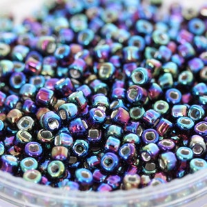 May include: A close-up view of a container filled with iridescent blue, purple, and green seed beads. The beads have a metallic finish and are arranged in a random pattern.