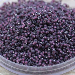 May include: A close-up shot of a large pile of small, dark purple seed beads. The beads have a matte finish and a small hole running through the center. They are scattered in a clear, shallow container, ready for crafting.