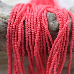 May include: A strand of coral pink seed beads. The beads are small and round, and they are strung together on a thin thread. The beads are arranged in a loose, flowing pattern.