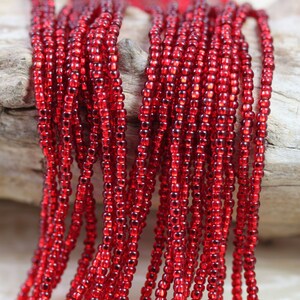 May include: A strand of red seed beads, approximately 100 strands, on a wooden surface. The beads are small and round, and they have a glossy finish.