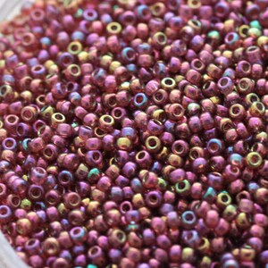 Puede incluir: Primer plano de un montón de pequeñas cuentas redondas. Las cuentas son principalmente de un color púrpura rojizo intenso con un brillo iridiscente, que refleja toques de verde, dorado y azul. Adecuado para la fabricación de joyas u otros proyectos de artesanía.