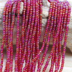 Pode incluir: Um fio de contas de sementes vermelhas iridescentes. As contas são pequenas e redondas, e têm um brilho metálico.