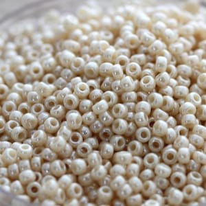 May include: A close-up image of a large quantity of small, round, off-white beads. The beads have a slight iridescent sheen.