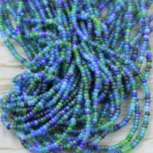 May include: A close-up of a pile of seed beads in shades of blue, green, and purple. The beads are strung together, creating a textured and colourful display. The beads are small and round, perfect for jewellery making or other crafts.
