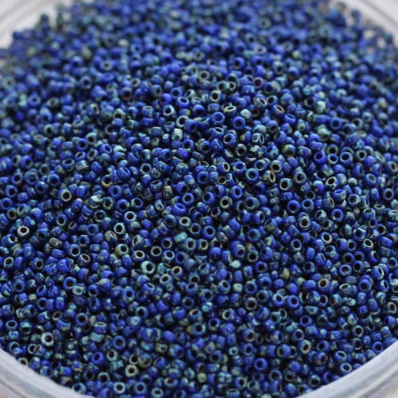 May include: A close-up view of a large quantity of small, round, blue glass beads. The beads have a slightly iridescent finish, giving them a subtle sparkle.