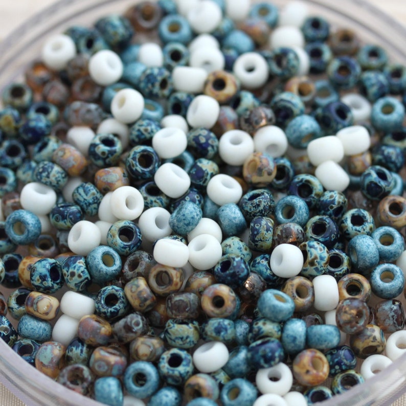 May include: A close-up of a bowl filled with small, round beads. The beads are a mix of white, blue, and brown colors. The beads have a speckled or marbled appearance.