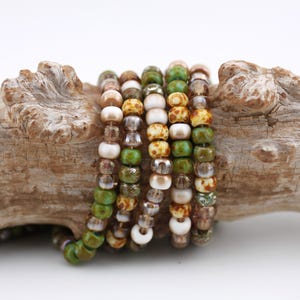 May include: Several beaded bracelets in shades of green, brown, white, and gold. The bracelets are arranged on a piece of driftwood, highlighting the natural colours of the beads and the wood.