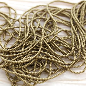 May include: A close-up of a pile of gold-colored seed beads. The beads are small and round, and they are arranged in a random pattern.