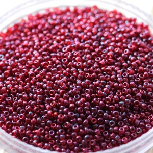 May include: A close-up shot of a clear container filled with numerous small, dark red seed beads. The beads are densely packed, creating a textured surface. The beads are uniform in size and shape, with a glossy finish, ideal for jewelry making and crafts.