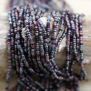 May include: A strand of seed beads in shades of purple, silver, and brown. The beads are small and round, and they are arranged in a random pattern.