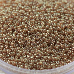 Puede incluir: Una vista de cerca de un contenedor lleno de pequeñas cuentas de semillas redondas de color marrón iridiscente. Las cuentas tienen un brillo sutil y son perfectas para la elaboración de joyas.