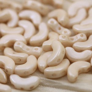 May include: A close-up of a pile of off-white ceramic beads shaped like crescent moons. The beads have a smooth, glossy finish.