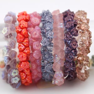 Puede incluir: Una colección de ocho hebras de cuentas de vidrio, cada hebra con un color y un diseño diferentes. Las cuentas son todas pequeñas y tienen forma de flor. Los colores incluyen morado claro, rojo, rosa, azul, rosa claro, morado, marrón y marrón claro.