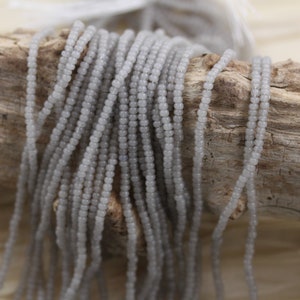 May include: A strand of light gray seed beads, arranged in rows, on a wooden surface.