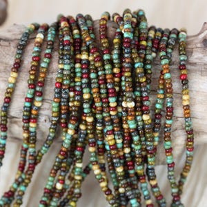 May include: A collection of small seed bead strands in various colours, including red, green, brown, and turquoise. The beads are strung together, creating a textured effect. The beads are resting on a piece of driftwood.
