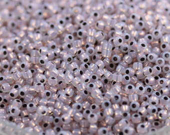 May include: A close-up view of a large quantity of small, round, iridescent white beads with black centers. The beads are arranged in a random pattern and appear to be made of glass.