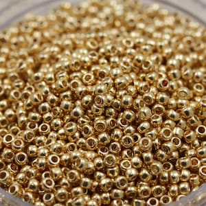 May include: Close-up of a pile of small, shiny, gold-colored beads. The beads are round with a hole through the center, and they reflect light, creating a sparkling effect. These beads are likely used for jewelry making or other craft projects.