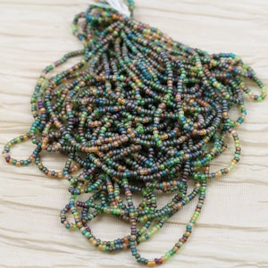 May include: A pile of small, iridescent glass beads in shades of green, brown, and gold. The beads are arranged in a loose, flowing pattern, creating a visually appealing display.