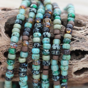 May include: Close-up of a strand of turquoise, green, brown, and black seed beads. The beads are arranged in a row and are all different sizes and shapes.