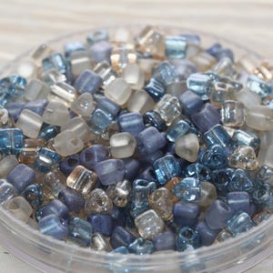 May include: A collection of small, irregular glass beads in shades of blue, gray, and translucent beige. The beads feature a mix of matte and glossy finishes, with some appearing clear and others opaque, all contained within a circular container.