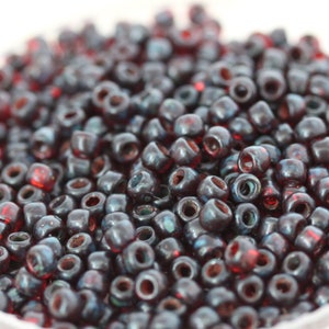 May include: A close-up of a pile of dark red and blue glass seed beads. The beads are small and round, with a slightly iridescent finish.