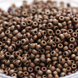 May include: A close-up view of a pile of small, round, brown seed beads. The beads have a metallic sheen and are arranged in a random pattern.
