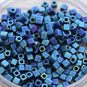 May include: A collection of small, square beads in shades of blue and teal. Each bead has a square hole running through the center. The beads are scattered in a clear container, creating a textured, iridescent appearance.