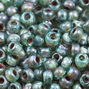 May include: A close-up of a pile of small, round, iridescent green glass beads with a red-brown hole in the center. The beads have a speckled, textured surface.
