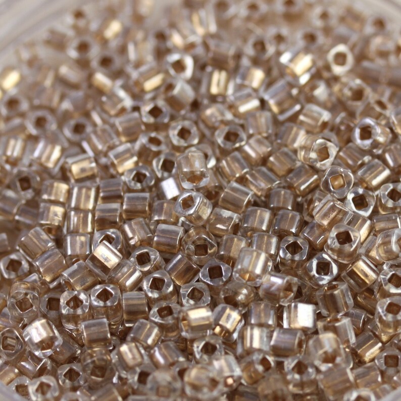 May include: A close-up of a large quantity of small, clear glass beads with a gold iridescent finish. The beads are in a round container.