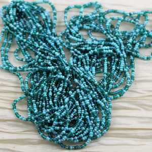May include: A strand of turquoise and teal seed beads, with iridescent shimmer, in a loose pile on a white fabric background.