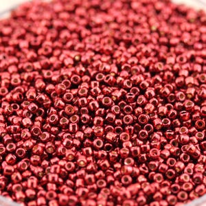 May include: Close-up of a pile of small, shiny, red seed beads. The beads are densely packed together, filling the frame. The beads have a metallic sheen, reflecting light. These beads are commonly used for jewelry making and crafting.