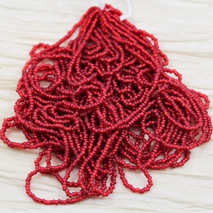 May include: A collection of small, red seed beads strung together. The beads are a deep, rich red color and appear to be made of glass or a similar material. The strands are bundled together, with a white string at the top.
