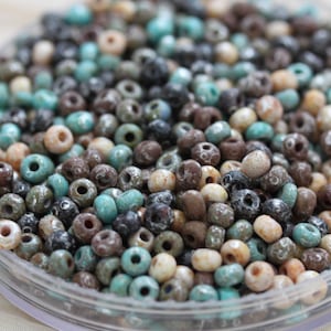 May include: A collection of small, round beads in various colors, including turquoise, brown, cream, and black. The beads have a matte finish and are scattered in a clear container. These beads are suitable for jewelry making and crafts.