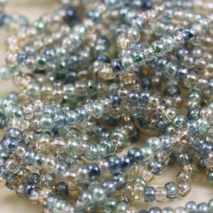 May include: A close-up of a pile of small, round glass beads in shades of blue, green, and beige. The beads are arranged in a random pattern, creating a textured and colorful surface.