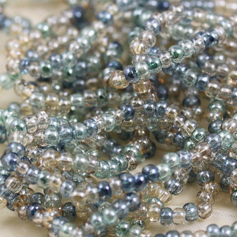 May include: A close-up of a pile of small, round glass beads in shades of blue, green, and beige. The beads are arranged in a random pattern, creating a textured and colorful surface.