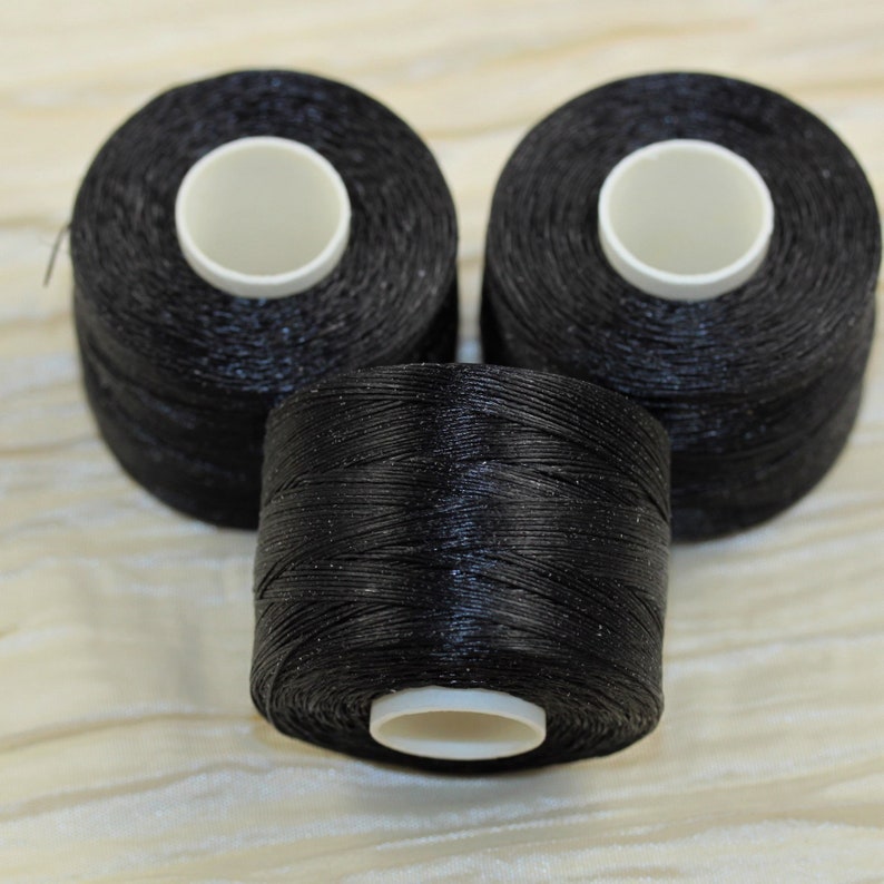 Puede incluir: Tres carretes de hilo negro con un ligero brillo. El hilo est&aacute; enrollado apretado en cada carrete.