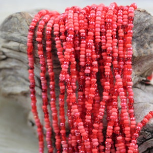 Puede incluir: Primer plano de una hebra de cuentas de semillas rojas. Las cuentas son pequeñas y redondas, y tienen un ligero brillo. Las cuentas están enhebradas en un hilo transparente, y están dispuestas en un patrón suelto y fluido.