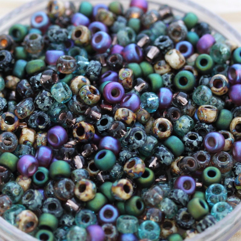 K&ouml;nnte beinhalten: Eine Mischung aus kleinen, bunten Glasperlen in den Farben T&uuml;rkis, Gr&uuml;n, Lila und Braun. Die Perlen haben verschiedene Oberfl&auml;chen, darunter irisierend, matt und gesprenkelt.