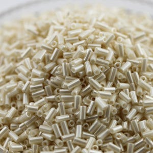 Op de afbeelding: Een close-up van een stapel ivoorkleurige, cilindrische kralen. Elke kraal is ongeveer 0,6 cm lang en heeft een glanzende, parelmoerachtige afwerking. De kralen zijn verspreid, waardoor een textuur ontstaat, ideaal voor sieraden maken en knutselen.