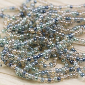 May include: A close-up of a pile of small, round, glass beads in shades of blue, green, and beige. The beads are translucent and have a slight iridescent sheen.