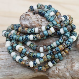 May include: A close-up of a strand of blue, white, and brown glass beads. The beads are small and round, and they have a speckled pattern. The beads are arranged in a coil on a piece of driftwood.