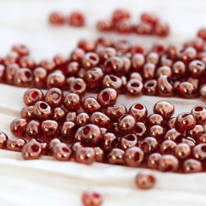 Op de afbeelding: Een grote stapel kleine, ronde, koperkleurige glazen kralen. De kralen hebben een glanzende, metallic afwerking.