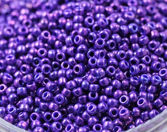 May include: A close-up of a container filled with small, round, purple beads. The beads have a metallic sheen and are arranged in a random pattern.
