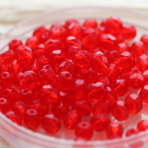 Puede incluir: Primer plano de numerosas cuentas rojas facetadas. Las cuentas son translúcidas y reflejan la luz, creando un efecto brillante. Están contenidas en un plato claro y poco profundo, probablemente para manualidades o bisutería. Las cuentas miden aproximadamente 0,5 cm de diámetro.